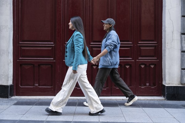 A Latin lesbian couple walks hand in hand along a city street. Their casual attire reflects modern urban style. They smile, enjoying a peaceful moment together