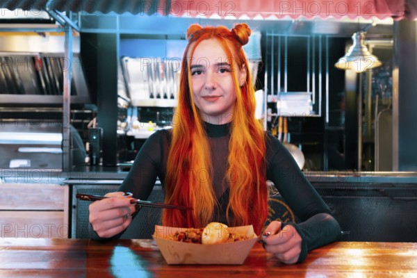 A woman with vibrant orange hair eats using chopsticks at an Asian food market She is seated at a wooden table, showcasing culinary culture and enjoyment of diverse flavors