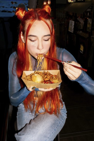A woman with vibrant hair savoring noodles with chopsticks in an Asian food market, capturing the essence of culinary exploration and cultural diversity