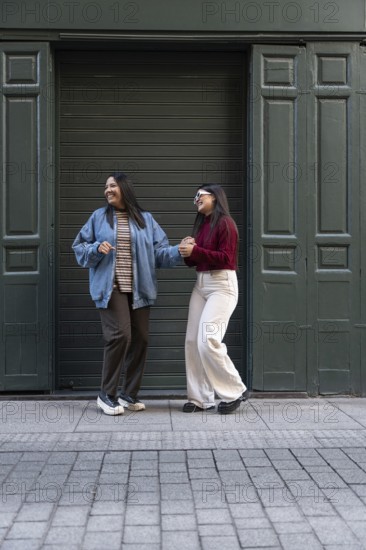 A cheerful latin lesbian couple, wearing casual outfits, holds hands while enjoying a fun moment outside in front of a dark green garage door. Their expressions radiate joy and connection