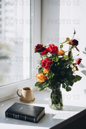 A serene windowsill featuring a bouquet of vibrant roses in a glass vase, a Bible, and a warm-toned mug, bathed in natural sunlight A peaceful and contemplative scene