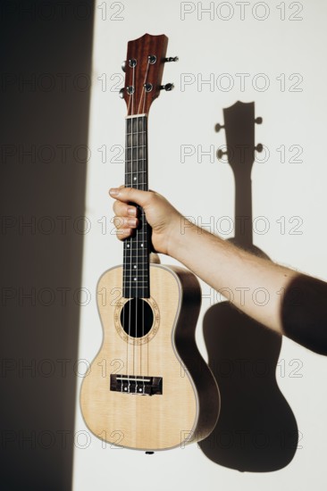 A person holds a ukulele against a plain wall, with sunlight casting a clear shadow The warm tones and simple backdrop emphasize the instrument's details
