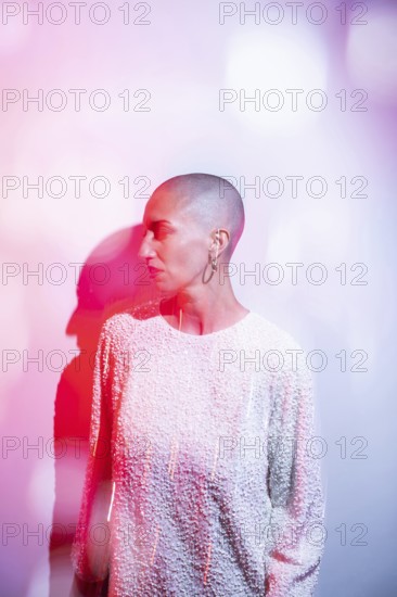 A middle-aged woman with a shaved head, wearing a textured white top, is illuminated by artistic lighting. The ethereal pink tones add a dreamlike quality to the scene