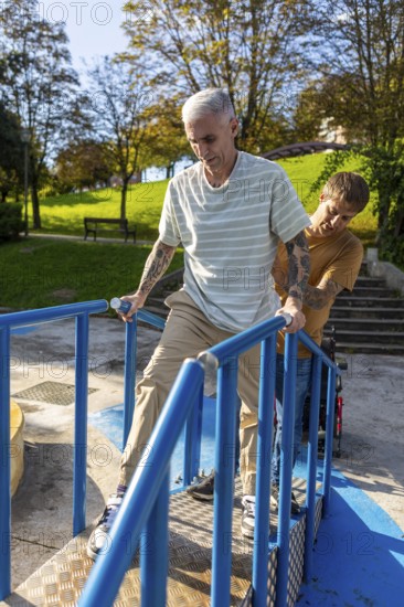 A man with tattoos, holding rails, is assisted by a friend on an accessible ramp in a park Sunlight filters through trees, illuminating the supportive interaction