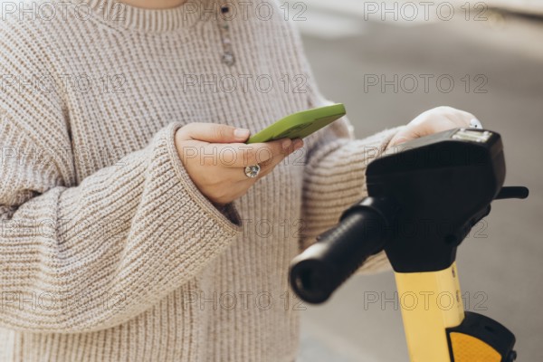 Cropped unrecognizable woman scans QR code with a smartphone to rent an electric scooter. The focus is on her hands holding both the phone and scooter's handle