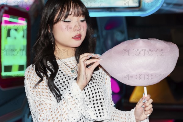 Asian Gen Z girl enjoys cotton candy in a vibrant arcade setting The colorful lights illuminate the playful atmosphere, capturing youthful joy and modern fun