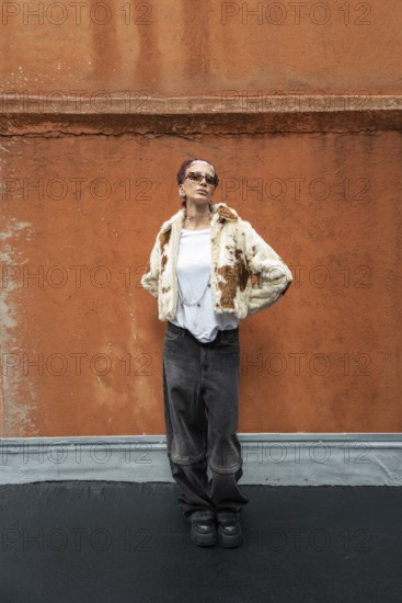 Stylish woman posing in a trendy fur jacket and sunglasses, set against a textured orange wall. The outfit combines casual and chic elements for a modern look