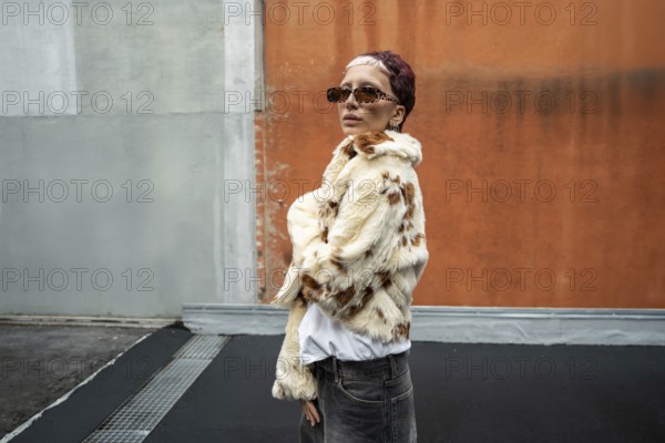 Fashionable woman in stylish faux fur jacket and sunglasses poses confidently against an urban background featuring gray and orange walls. The outdoor scene exudes modern elegance