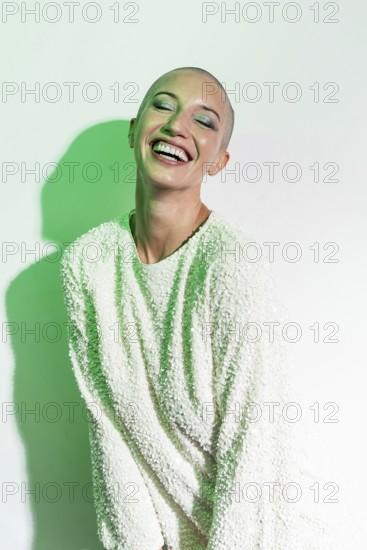 A cheerful middle-aged woman with a shaved head smiles brightly. She wears a textured white sweater, standing against a light background with green lighting