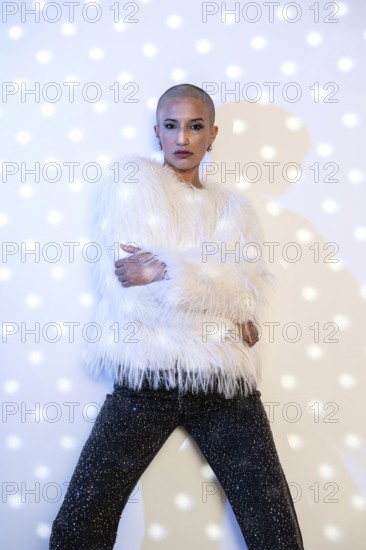 A middle-aged woman with a shaved head poses confidently. She wears a fur-like white jacket and glittery black pants, standing against a dotted light backdrop