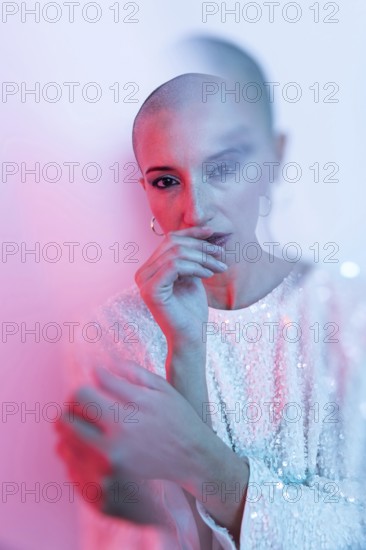 A striking image of a middle-aged woman with a shaved head, captured in a soft pink and blue light. She wears a sequined top, evoking elegance and confidence