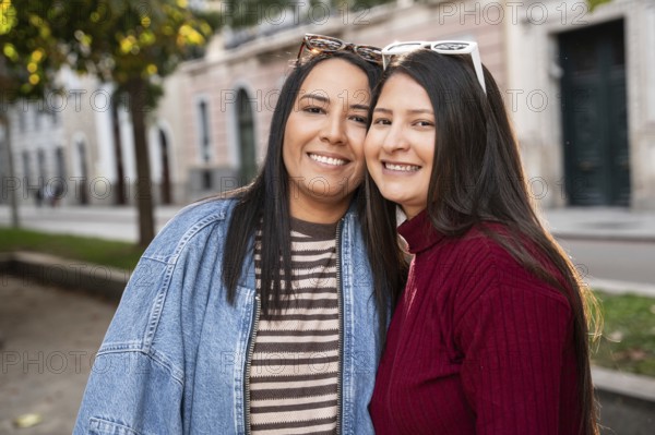 Portrait of a joyful Latin lesbian couple looking at camera against city background on a sunny day while smiling broadly