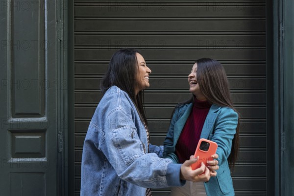 A smiling latin lesbian couple share a moment of joy outside, with one holding a smartphone. They are in front of a green door, embracing their time together in a casual setting
