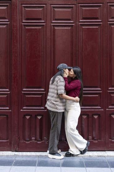 A loving latin lesbian couple shares a tender moment before a rich red wooden door. They embrace closely, symbolizing affection and unity, reflecting a romantic urban setting
