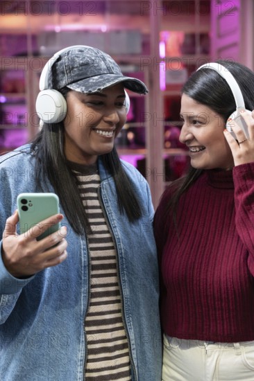 A joyful Latin couple shares a moment, smiling and listening to music with headphones. Their casual attire and cheerful expressions highlight a relaxed, warm interaction