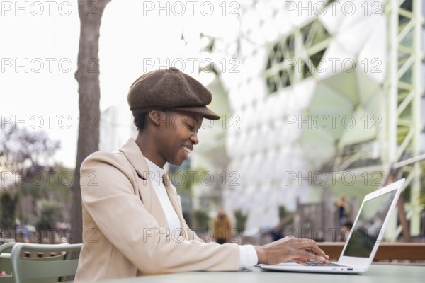 A black woman wearing a stylish hat and beige coat uses her laptop on an outdoor table in a modern urban environment, exuding confidence and sophistication
