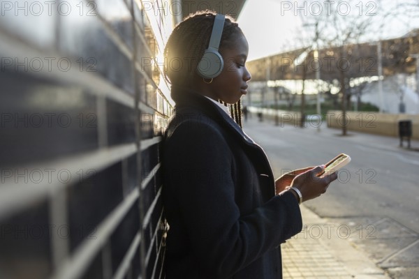 An African American businesswoman, sporting braids and headphones, is focused on her smartphone during a break outdoors, illuminated by soft sunset light