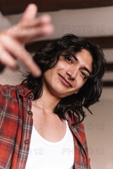 A smiling young man in a white tank top and plaid shirt extends his hand towards the camera, giving a friendly and inviting gesture in a warmly lit indoor setting