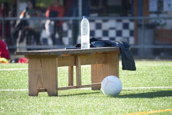 A wooden bench on a soccer field holds a water bottle and jacket, with a ball nearby. Sunlight casts shadows on the green artificial turf, creating a serene sports scene