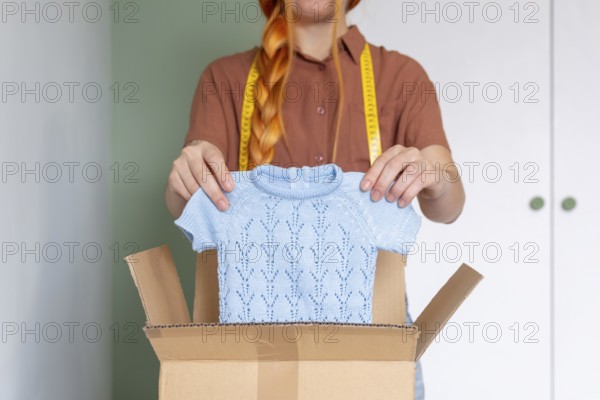 A woman is organizing her closet, preparing clothes for online sale. She holds a blue knit sweater over a cardboard box. A tape measure hangs around her neck
