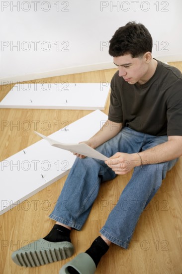 A young man sits on a wooden floor, reading assembly instructions for new furniture parts spread around him
