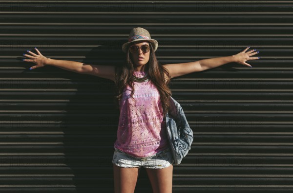 A fashionable woman poses confidently against a dark background, wearing a pink sleeveless top, shorts, a hat, and sunglasses, exuding summer style and confidence