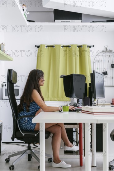 A young woman in a blue dress sits at a desk working on a computer in a modern home office. The setting includes a green curtain and books, creating a cozy workspace