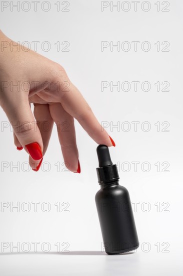 Cropped unrecognizable female hand with vibrant red manicure gracefully touching a sleek black cosmetic dropper bottle against a white background. Provides an elegant presentation of beauty packaging