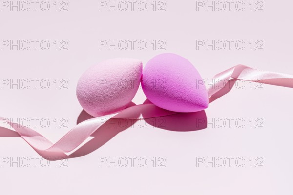 Two pink makeup sponges, also known as beauty blenders, intertwined with a soft pink ribbon, set against a pastel background