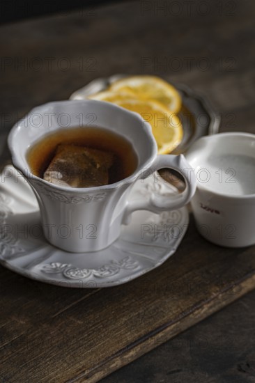 An elegant setting featuring a white porcelain tea cup with an embossed design, filled with English tea and a teabag. Accompanying the cup are fresh lemon slices on a saucer and a small milk jug