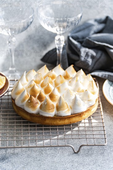 A beautifully baked a homemade lemon cake features toasted peaks, resting on a cooling rack. Crystal glasses and linen napkins create an elegant dessert setting
