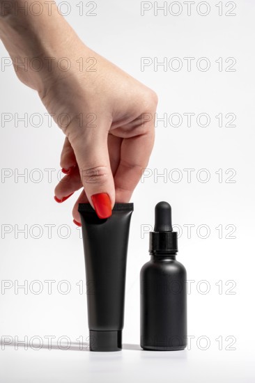 Cropped unrecognizable female hand with a bold red manicure holding a black tube and dropper bottle of cosmetics against a clean white background, showcasing elegance and style in beauty product presentation