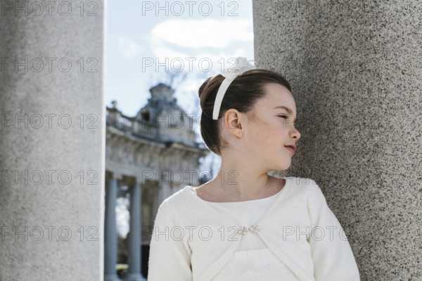 A young girl in a beautiful First Communion dress stands gracefully beside a large stone pillar, with a delicate headband adorning her hair. Sunlight filters through softly
