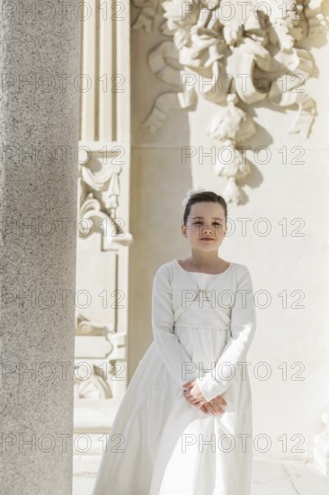 A young girl stands in her First Communion dress near an ornate architectural background, embodying grace and tradition on this special day