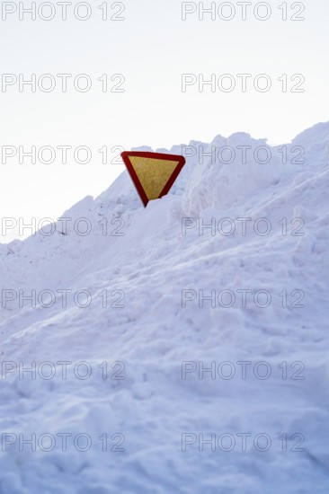 A yield sign partially buried in snow, standing amidst a snowy landscape in Swedish Lapland. The bright sign contrasts with the pure white snow under a clear sky