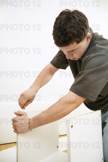 A focused young man assembles a new piece of white furniture using a screwdriver. He is seen inserting screws into the panels, showing hands-on skill in furniture assembly