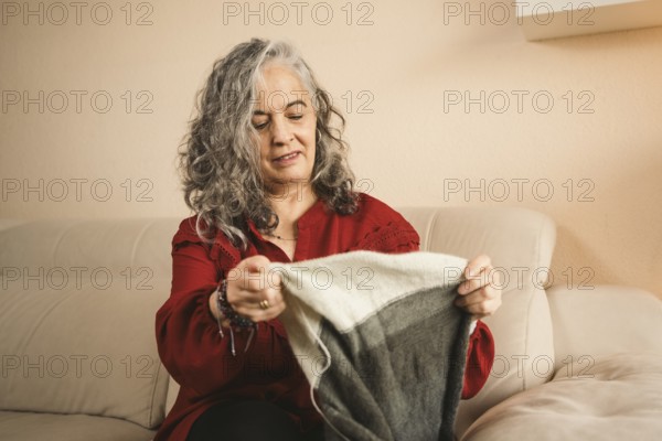 A woman in a red sweater, with gray hair, sits on a beige sofa knitting a gray and white piece. She is smiling and appears relaxed in her cozy living room setting