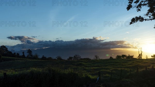 Capture the serene beauty of sunset over lush green fields under a vibrant sky lit by the descending sun, with misty mountains in the distance