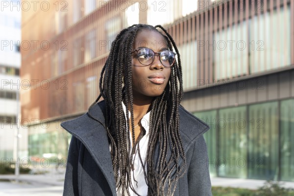 An African American businesswoman with braids and glasses takes a break outside a modern office building