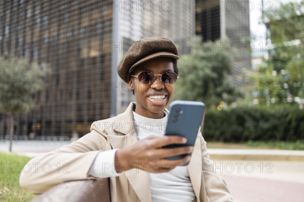 A stylish black woman wearing sunglasses and beret, sitting on a bench in the city She smiles while using her smartphone, embodying urban confidence and modern lifestyle