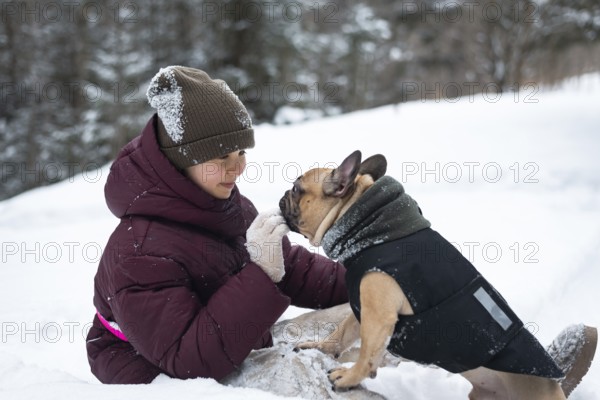 A heartwarming moment of a girl and their dog playing in the snow. The girl is bundled in winter clothing, sharing a joyful bond with a dressed-up dog amid a snowy landscape