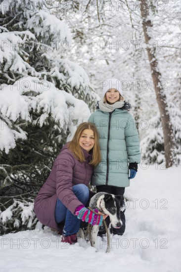 A joyful family poses with a dog in a snowy forest, surrounded by snow-covered trees. The bright winter attire contrasts with the serene white landscape
