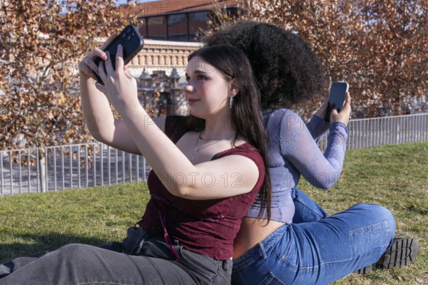 Two young women enjoy a sunny day outdoors while taking selfies. The lady on the left, with fair skin, captures a photo with her phone, and her friend with curly hair, identified as Hispanic or Latin, uses her device similarly