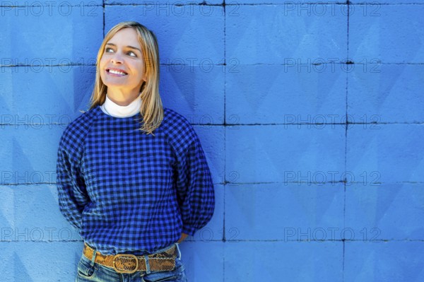 A cheerful woman smiles broadly while dressed in a blue checkered shirt and leaning against a vibrant blue wall. She looks upwards to her right, not directly at the camera