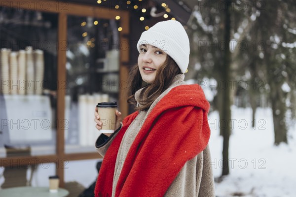 A young woman in a white beanie and red scarf smiles while holding a coffee cup, standing outdoors on a snowy day, with festive lights in the background