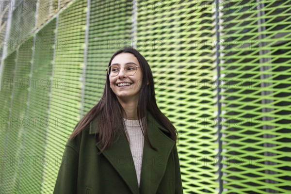 A woman in a stylish green coat smiles joyfully while standing against a vibrant, abstract green background Her glasses add sophistication to the modern urban setting