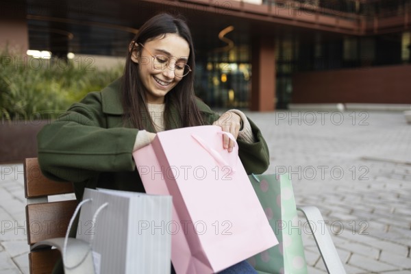 A young woman in a green coat sits outdoors, joyfully looking into a pink shopping bag She is surrounded by other colorful shopping bags, conveying excitement and leisure