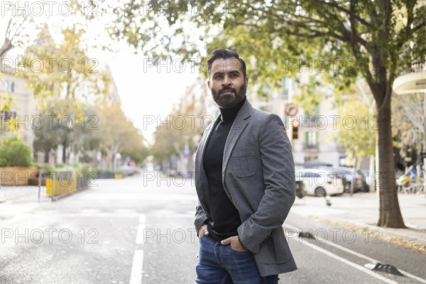 Stylish Indian businessman with a well-trimmed beard stands confidently on an urban street lined with trees, dressed in a sharp gray blazer and dark turtleneck, exuding a professional winter vibe