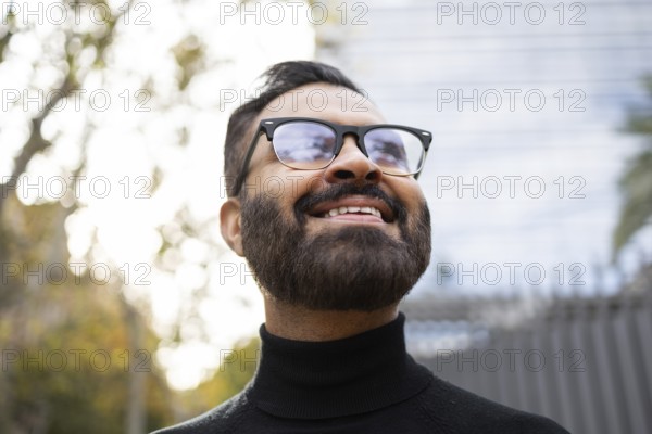 From below cheerful Indian businessman wearing sunglasses and a black turtleneck enjoys a sunny winter day outdoors, looking away from the camera with a joyful expression