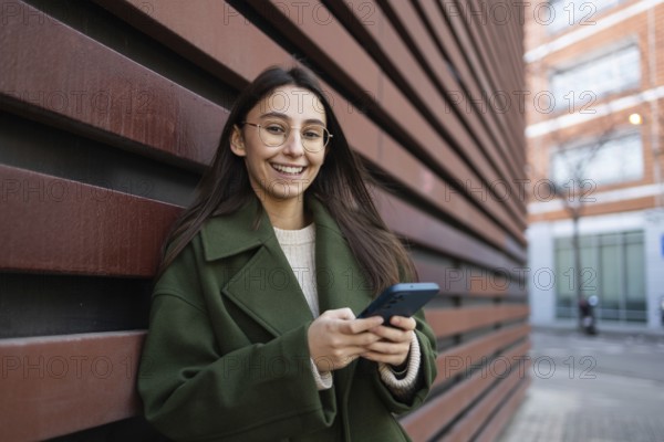 Young woman in a green coat smiling as she uses her smartphone, leaning against a red wall outdoors The urban backdrop adds a modern and vibrant feel to the scene